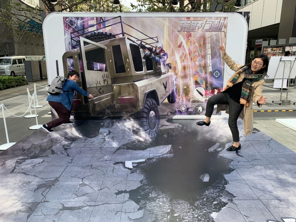 スマホ画面から車が飛び出す屋外イベント用トリックアート。ゲームのプロモーションで来場者がジャンプして撮影するフォトスポット